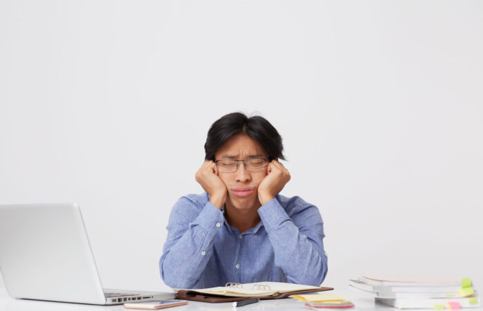 Tired sleepy asian young business man in glasses with head on hands sitting and sleeping at workplace at the table over white background